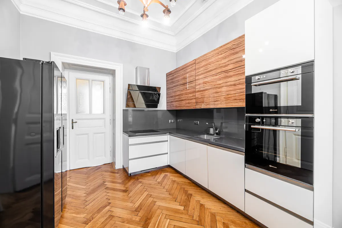 Modern kitchen with white cabinets, wood grain upper cabinets, and stainless steel appliances. Herringbone wood floors and gray walls.
