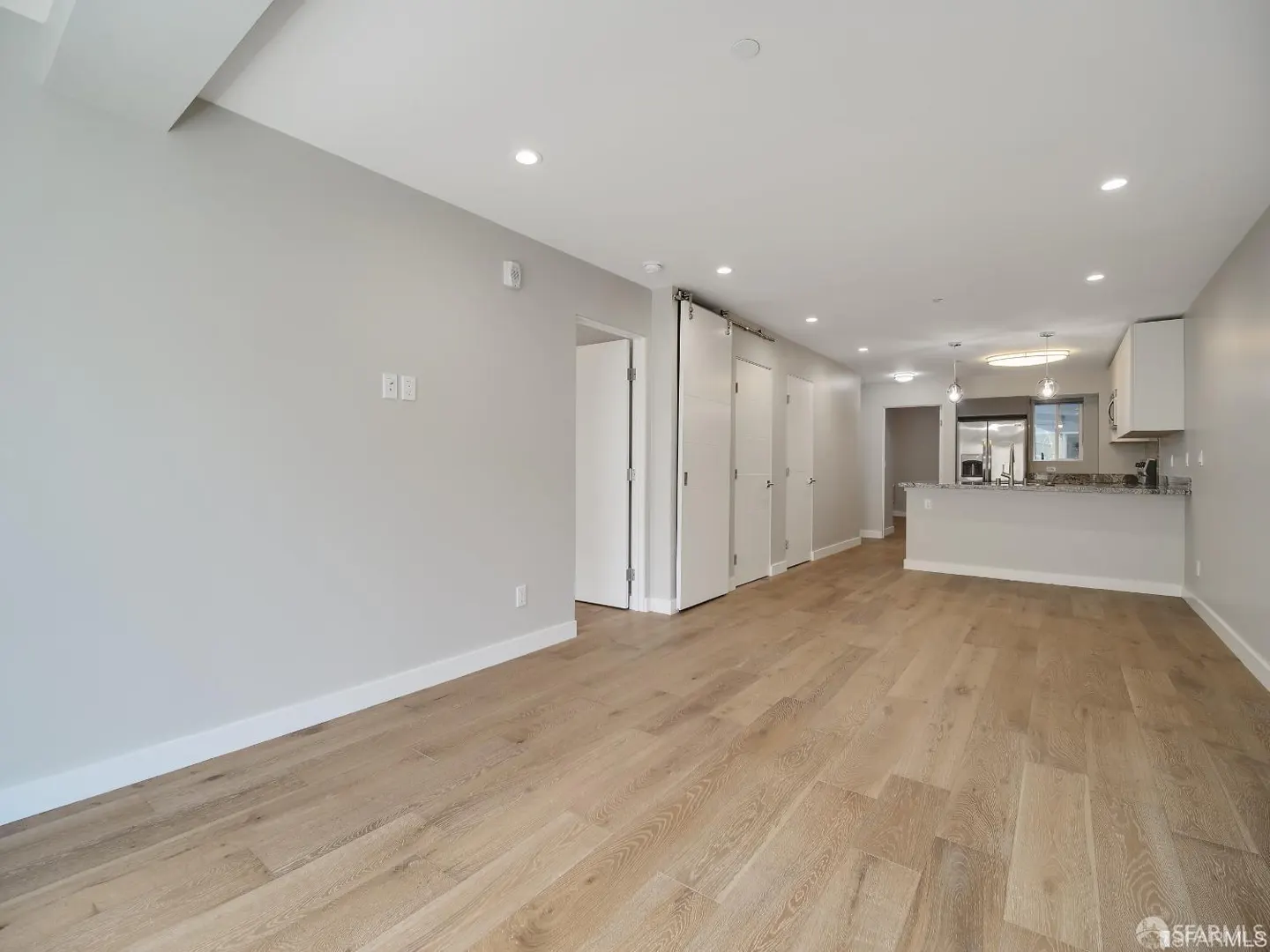 Bright, open-concept living space with light wood floors, gray walls, and white trim. White kitchen cabinets and stainless steel appliances are visible.