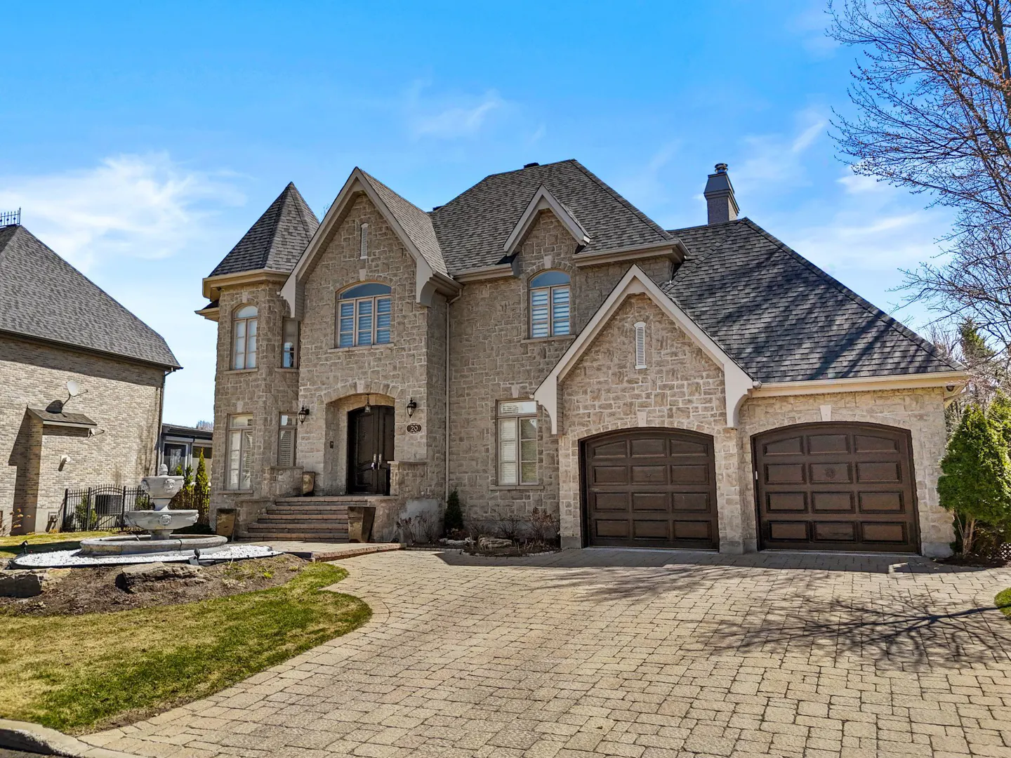 Exterior view of a large, two-story stone house with a gray roof and a brick driveway on a sunny day.