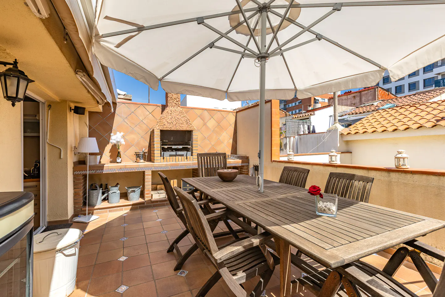Outdoor patio with a large wooden table and chairs under a white umbrella. A brick barbecue is in the background.