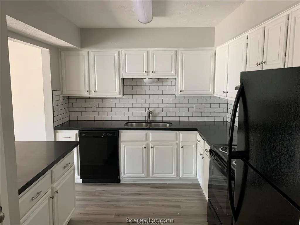 A kitchen with white cabinets, black countertops, and a black refrigerator. Subway tile backsplash and gray flooring.