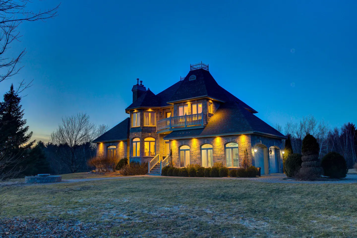 A large stone house with a dark roof is illuminated at dusk. The house has many windows and a balcony. A green lawn surrounds the house.