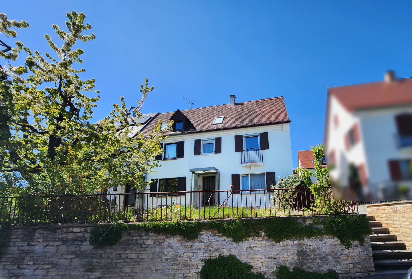 Two-story white house with brown shutters and a red tile roof, viewed from below a stone wall and iron fence.
