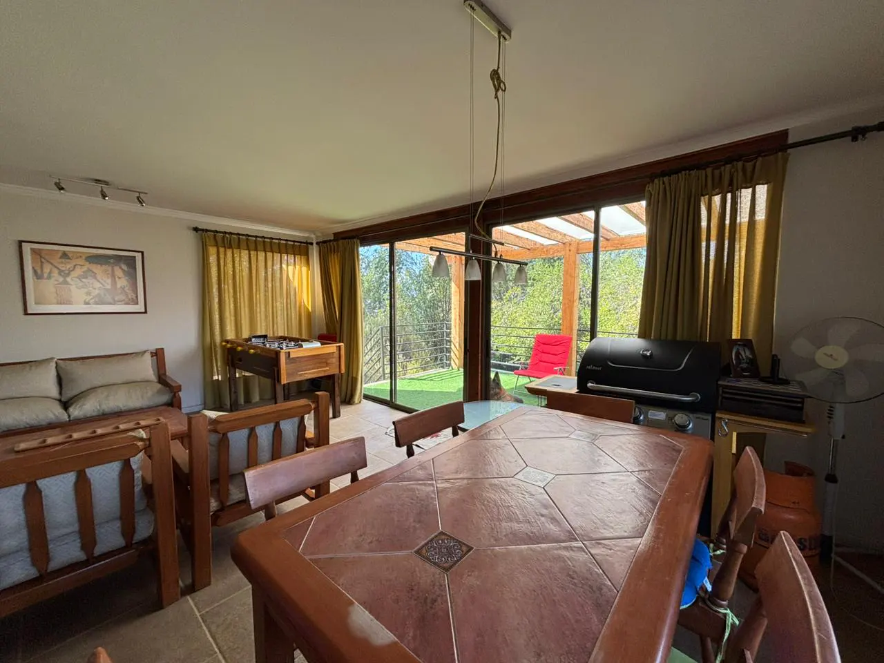 Living room with a large tile table, wooden chairs, sofa, and foosball table. A grill sits on the patio outside.