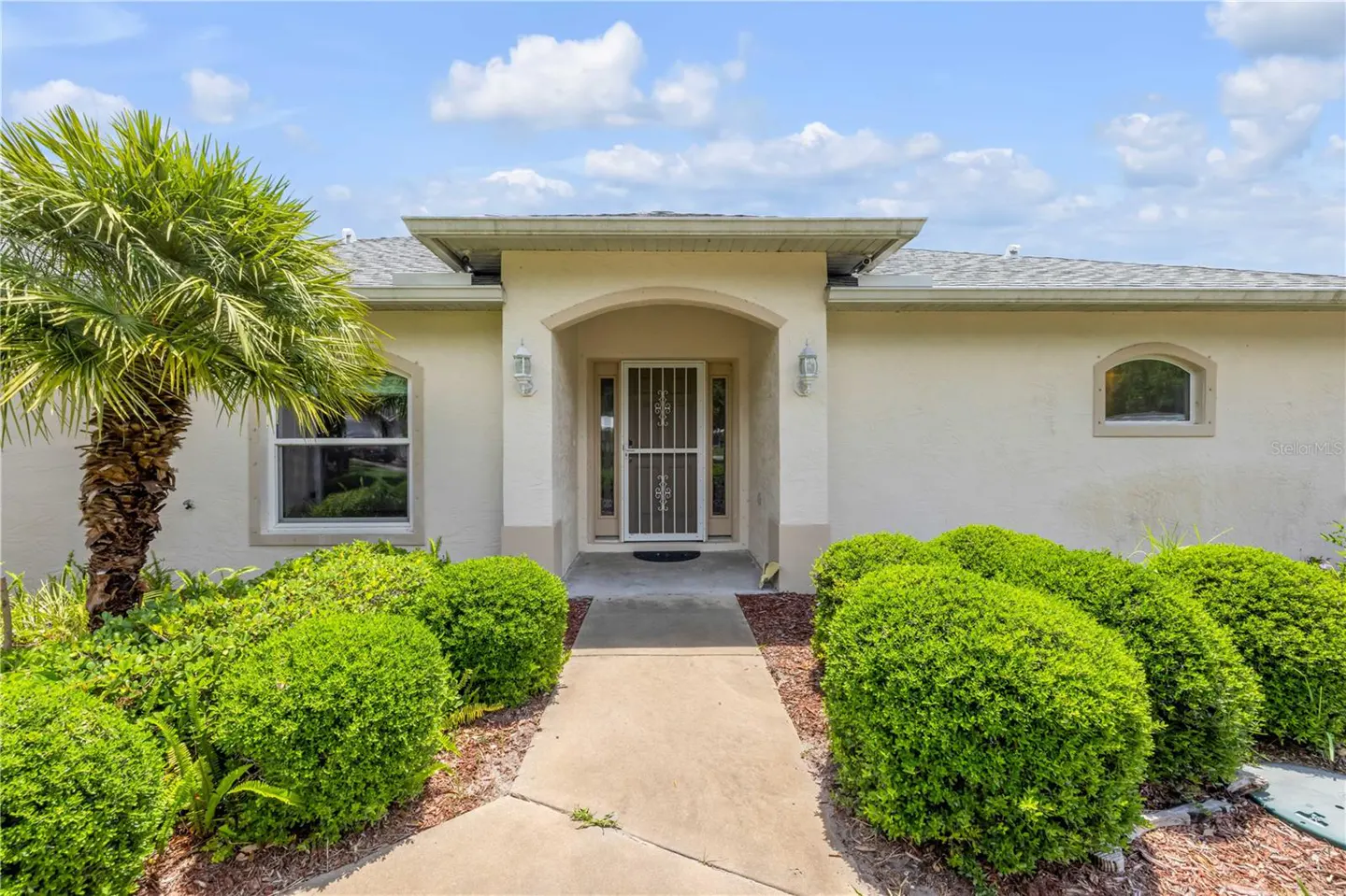 Beige single-story house with a gray roof, a palm tree on the left, and green bushes lining the walkway to the front door.
