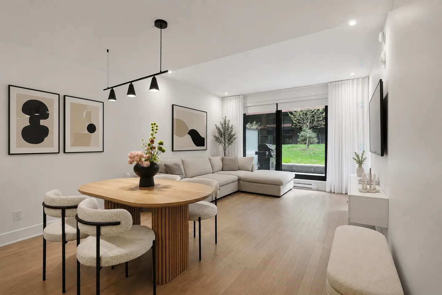 Bright, modern living space with wood floors, a round dining table with chairs, and a beige sectional sofa. Large windows overlook a green lawn.
