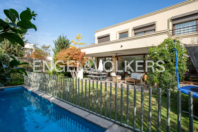 Backyard view of a two-story beige house with a patio, pool, and green fence under a blue sky.