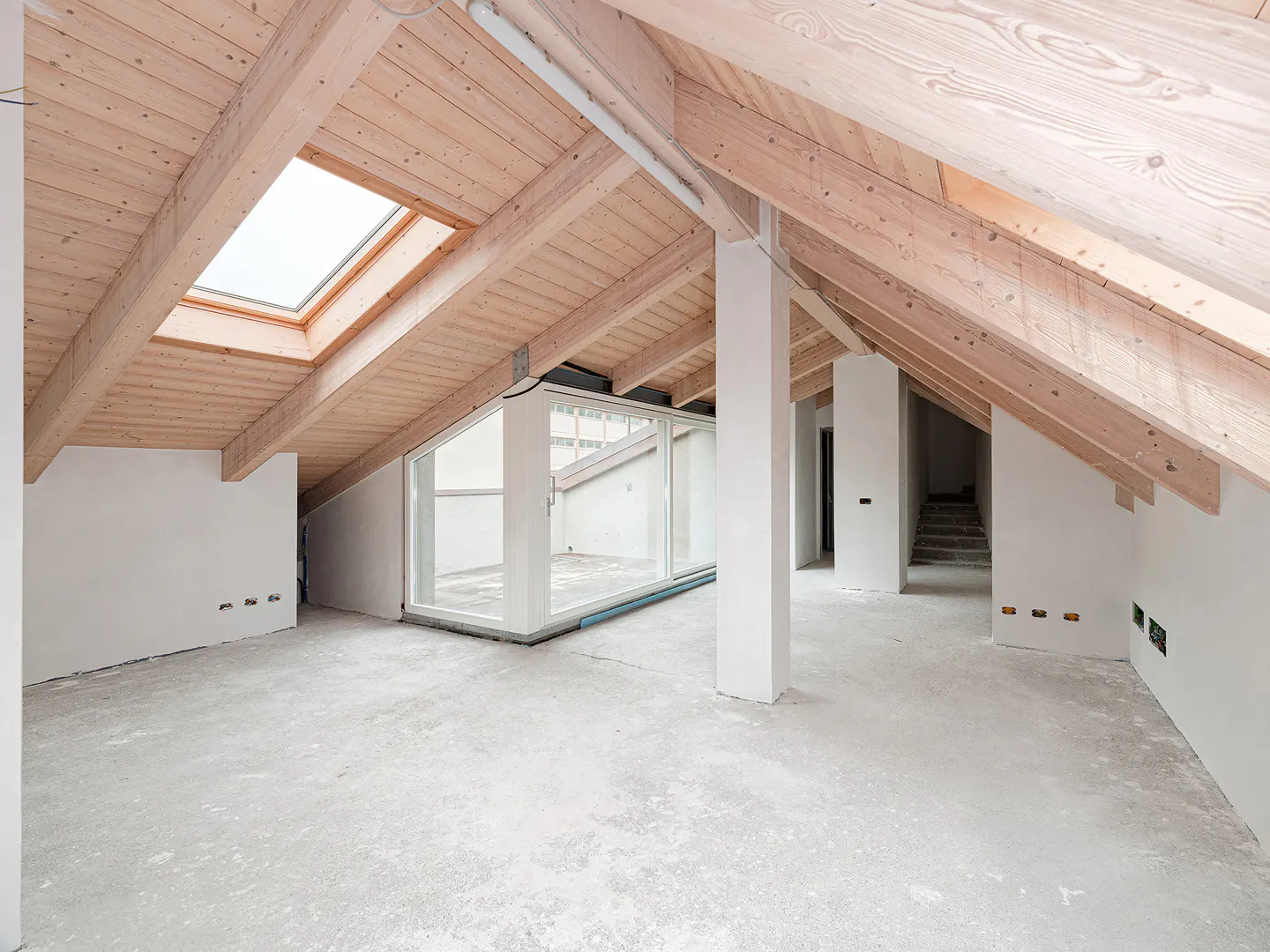 Unfinished attic space with exposed wood beams, a skylight, and white walls. Concrete floor and a glass-enclosed area are visible.