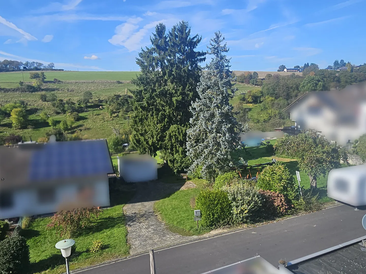 Elevated view of a suburban home with a paved driveway, lush green lawn, tall evergreen trees, and rolling hills beyond.