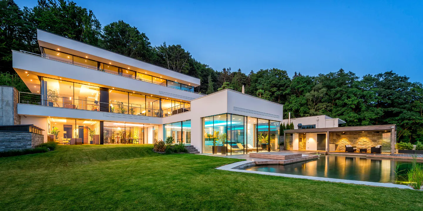 Modern white house with large windows, a pool, and green lawn at dusk. Trees in the background.