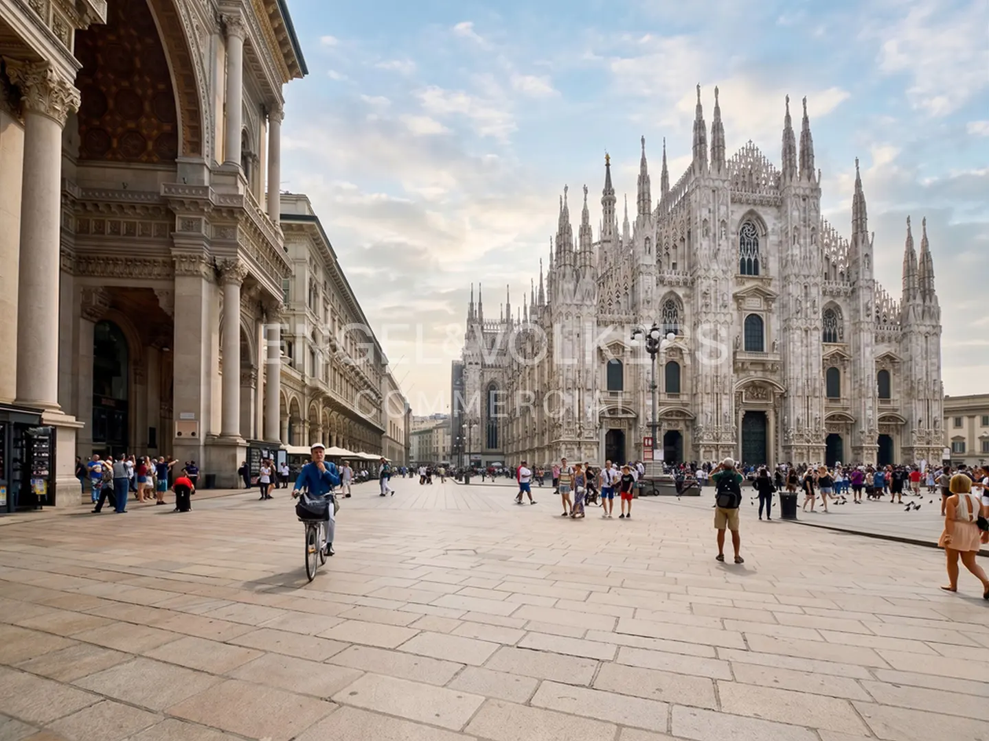 Milan's Piazza del Duomo with the Duomo cathedral in the background. People walk and a man rides a bike on the stone square.