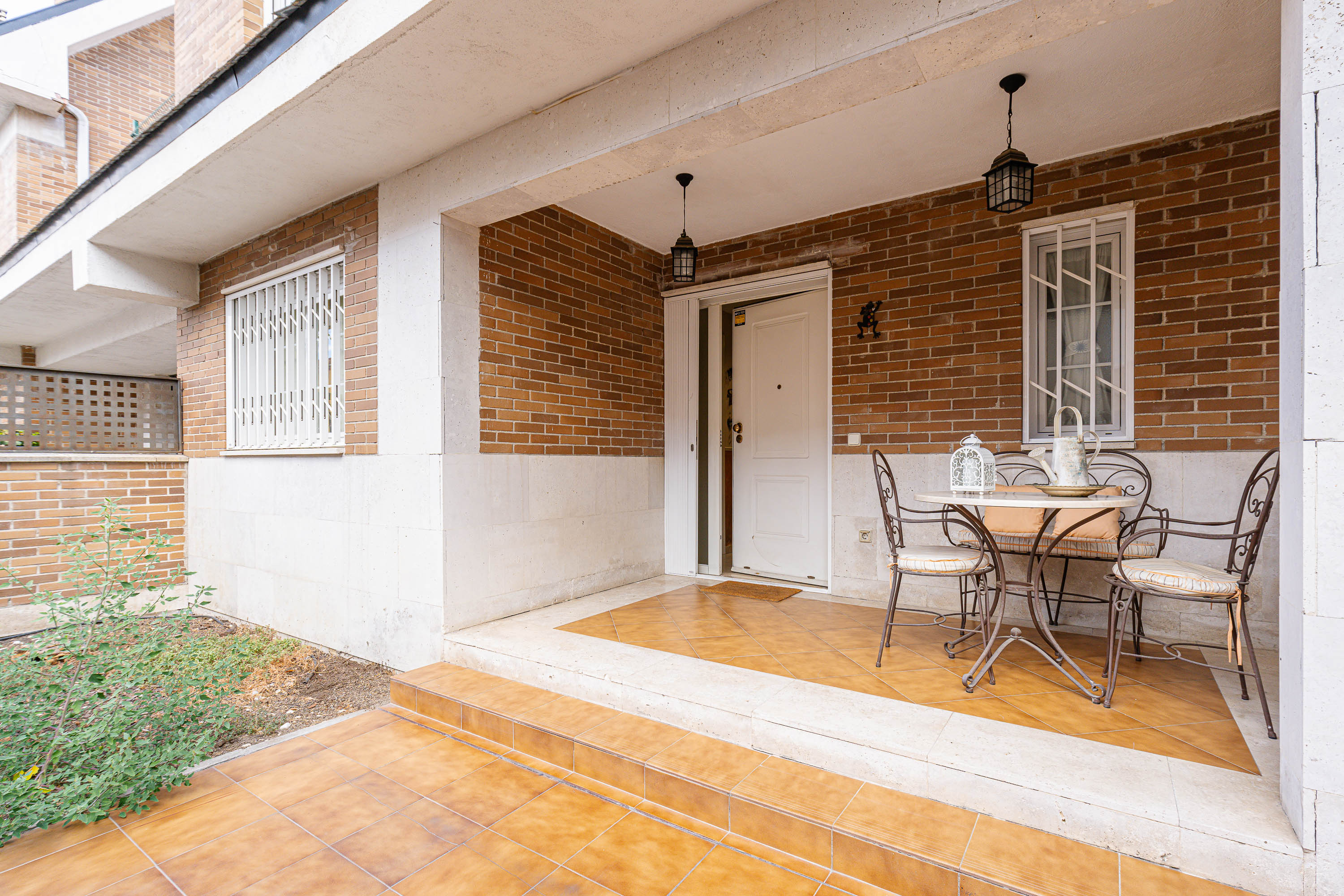A brick-walled porch with a white door, tiled floor, and a small bistro set with two chairs and a table. [AI-generated text]