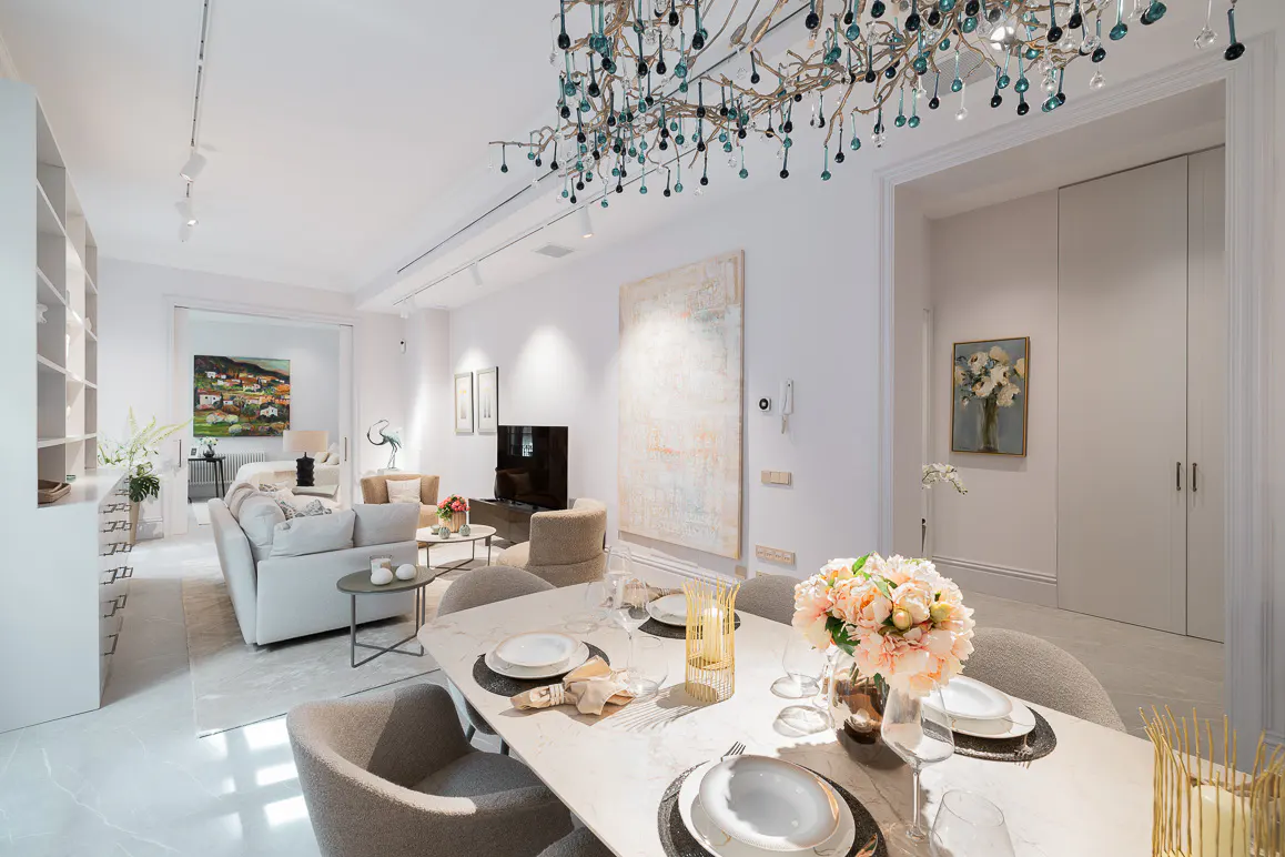 Bright, modern living and dining area with white walls, marble floors, and a unique chandelier. Table is set with flowers and tableware.