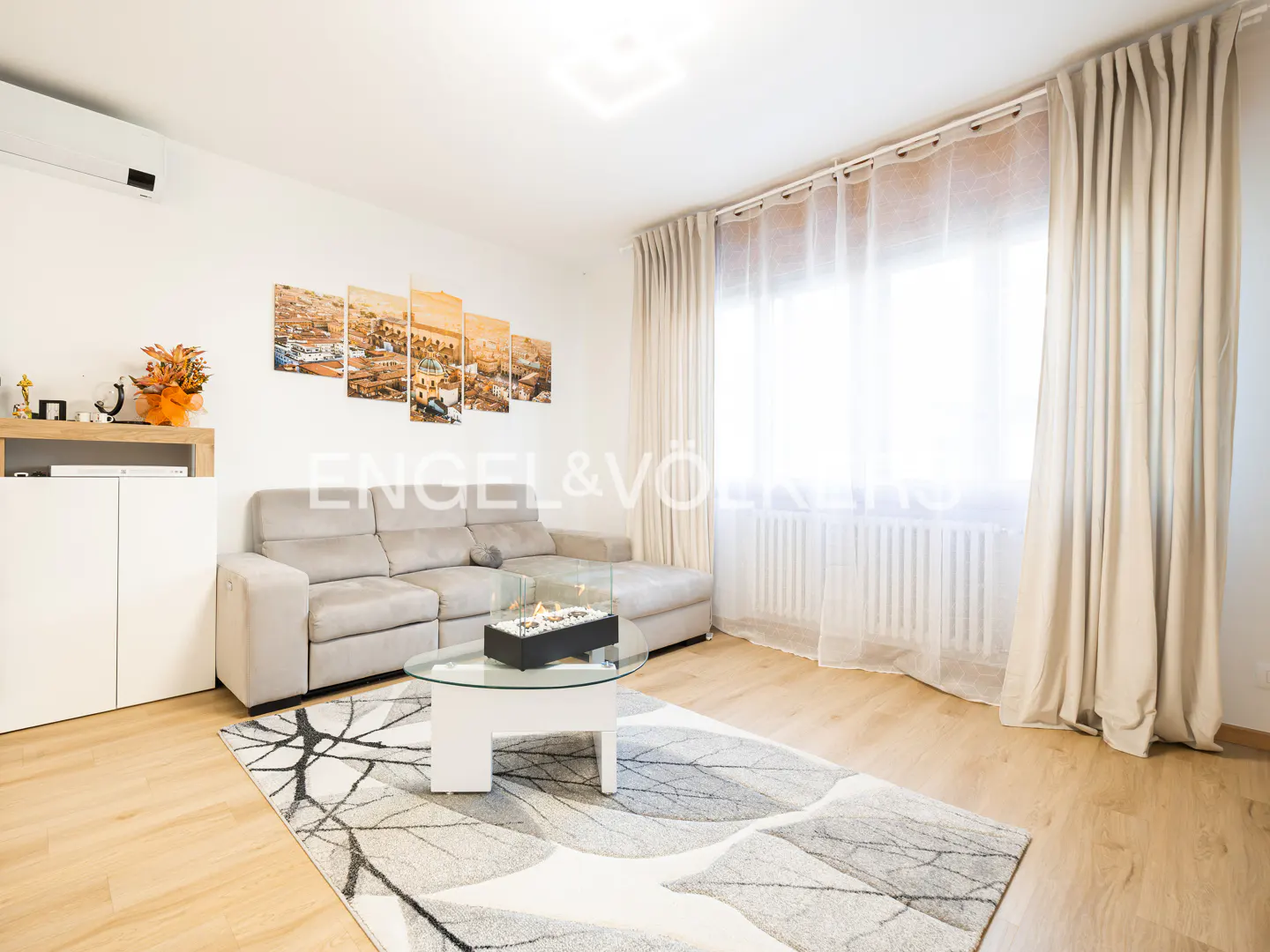Living room with a gray sectional sofa, glass coffee table, and a gray and white rug on a wood floor. Curtains cover a bright window.