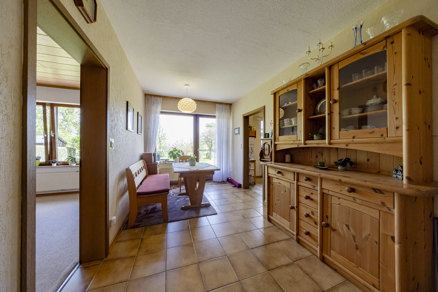 A dining room with a wooden hutch, table, and bench. The room has tile floors and a large window with a view of trees.