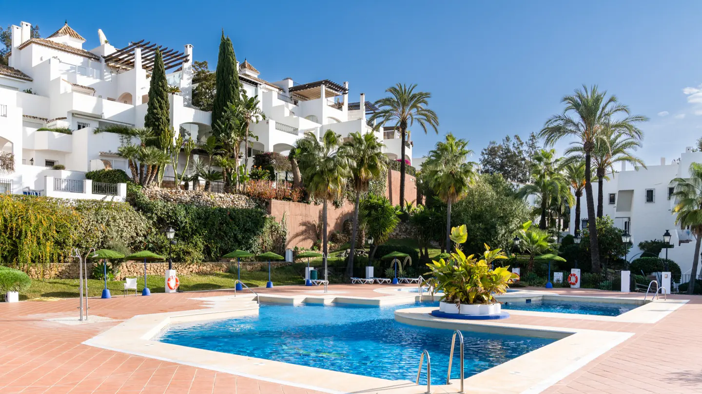 A multi-tiered pool with blue water and a red tile deck, surrounded by palm trees and white buildings under a clear blue sky.