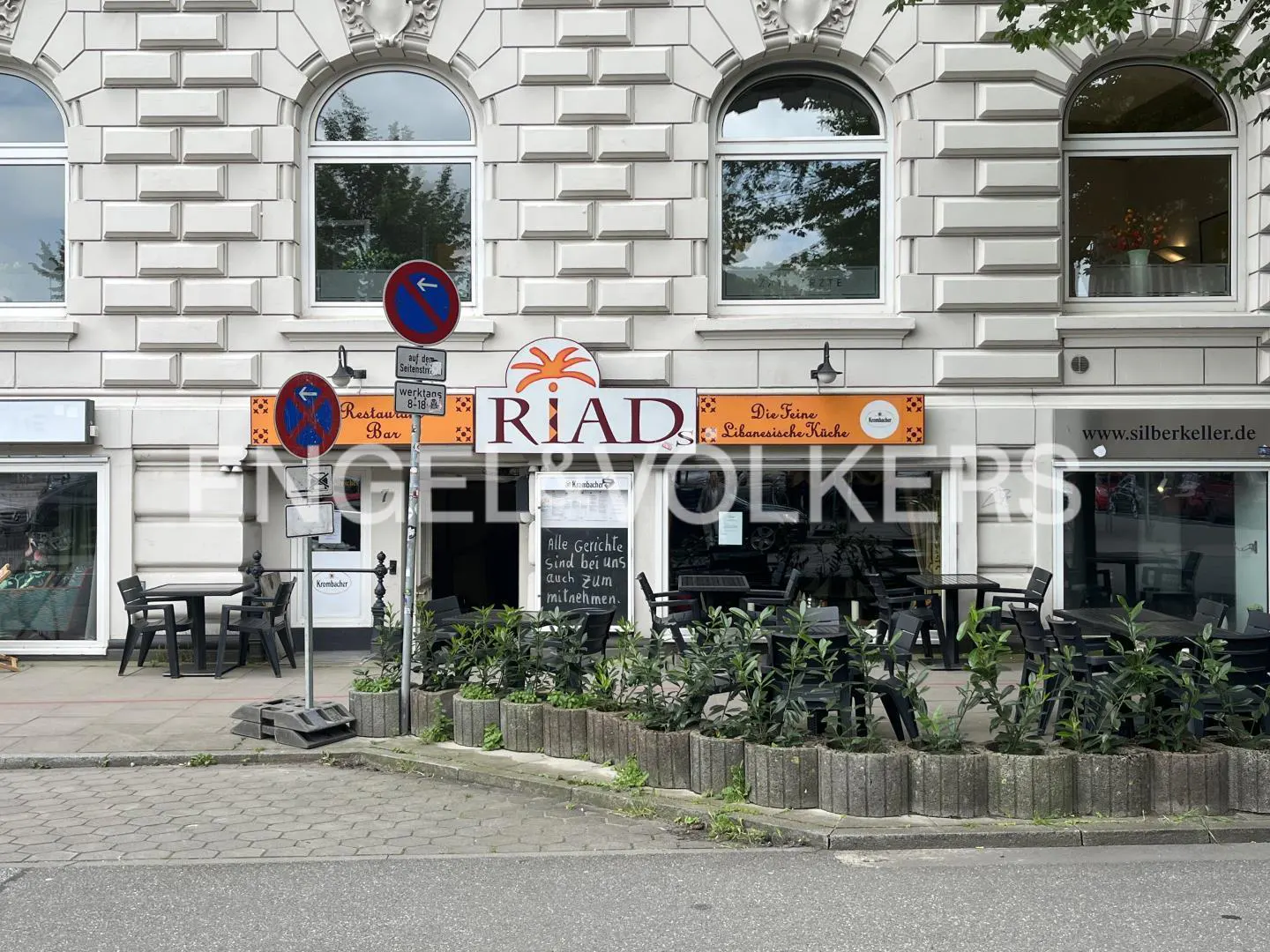 Exterior of "RIAD" restaurant with outdoor seating, signs, and a gray brick building facade.