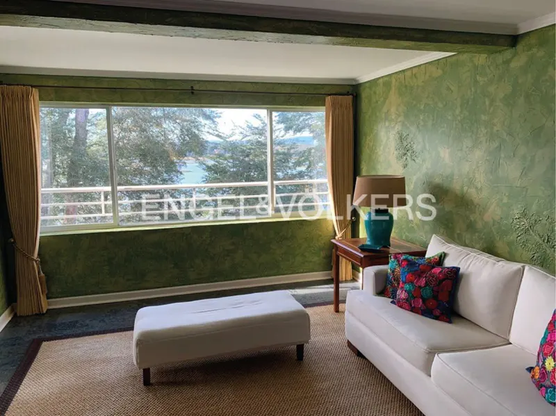 Living room with green textured walls, white sofa, ottoman, and a large window with a view of trees and water. A wooden table with a lamp sits beside the sofa.