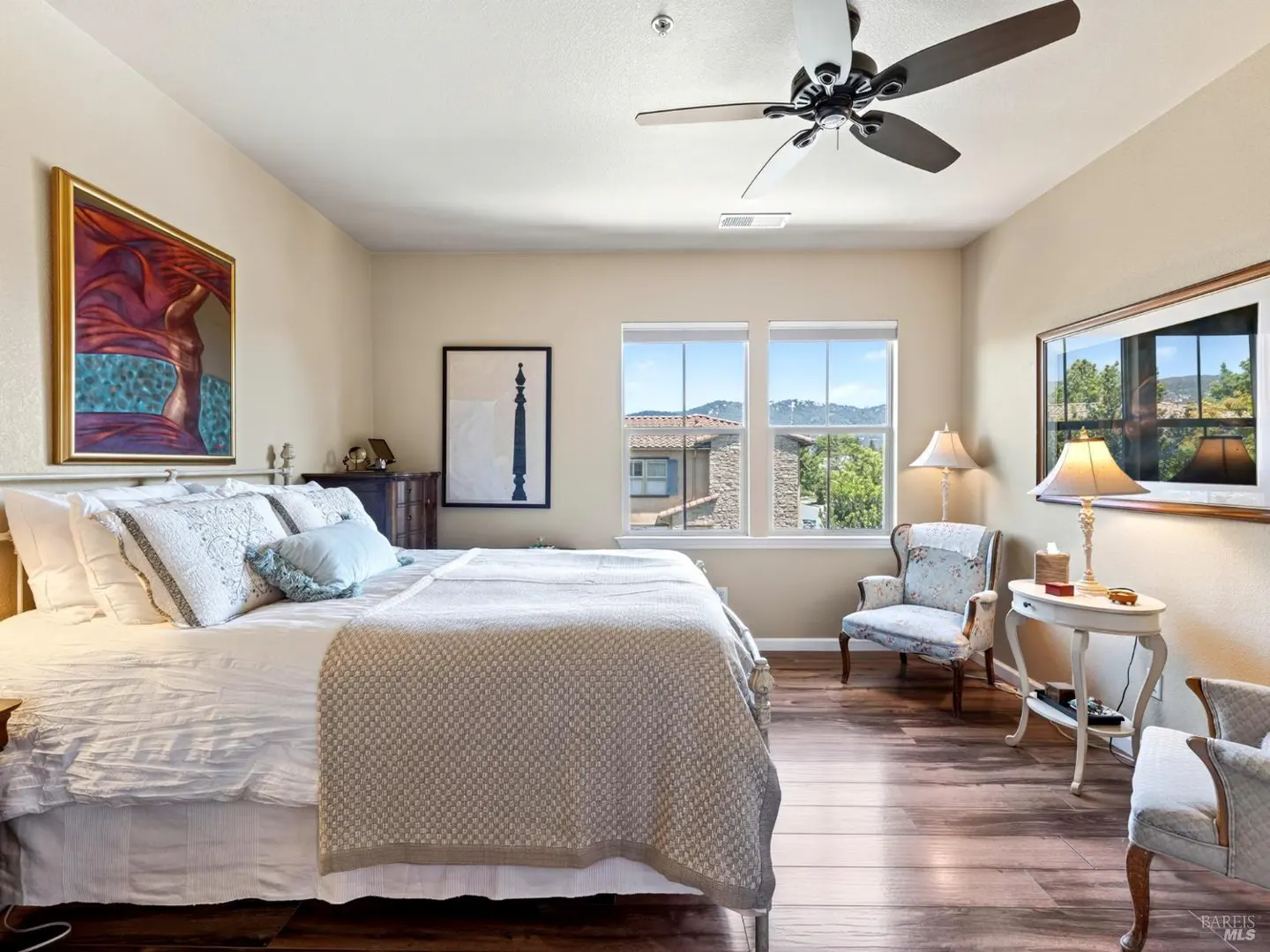 Spacious bedroom with a king bed, ceiling fan, two armchairs, a side table with a lamp, and windows overlooking hills.