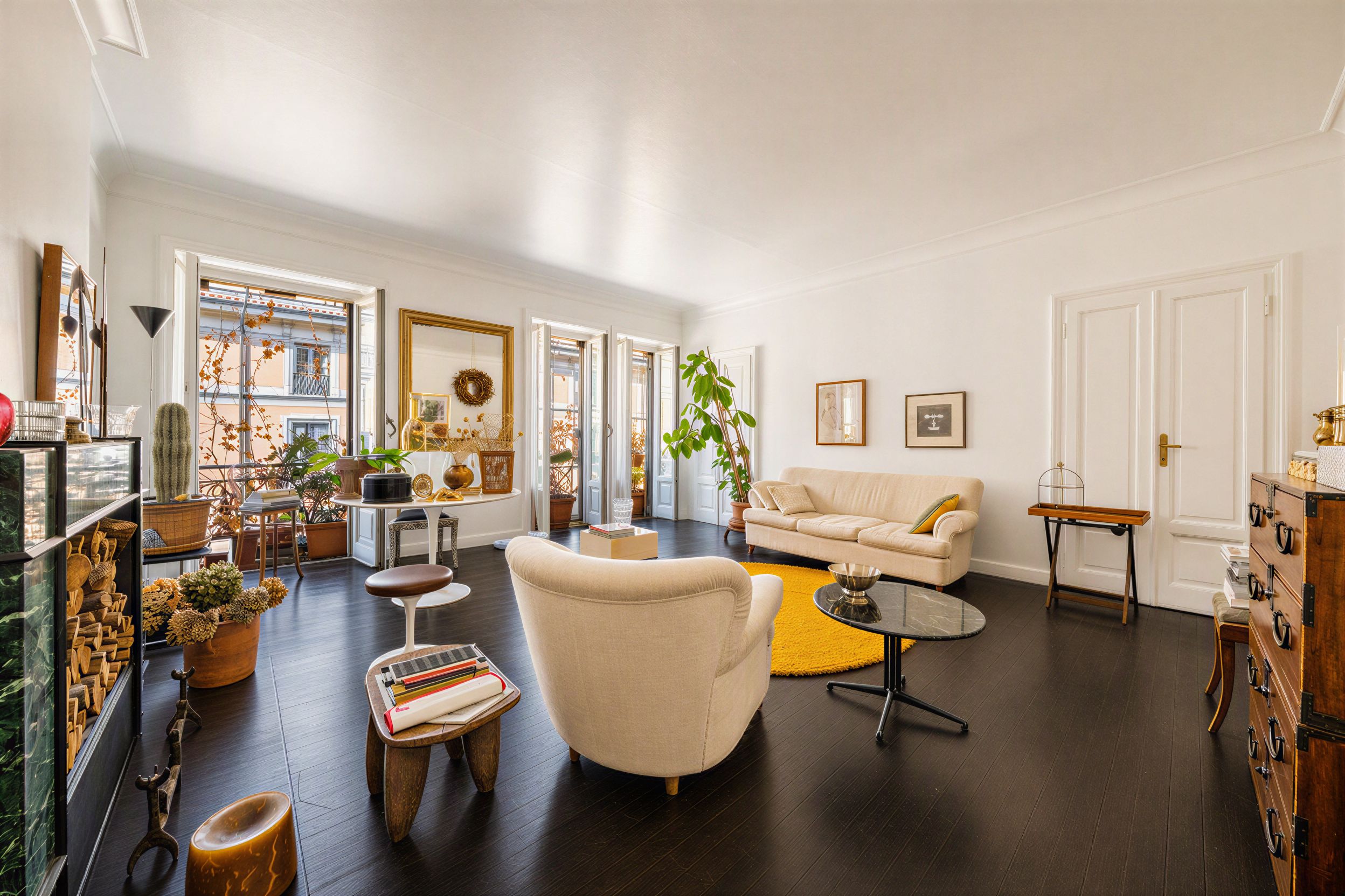Bright living room with dark wood floors, cream sofa and armchair, yellow rug, and large windows with balcony views.