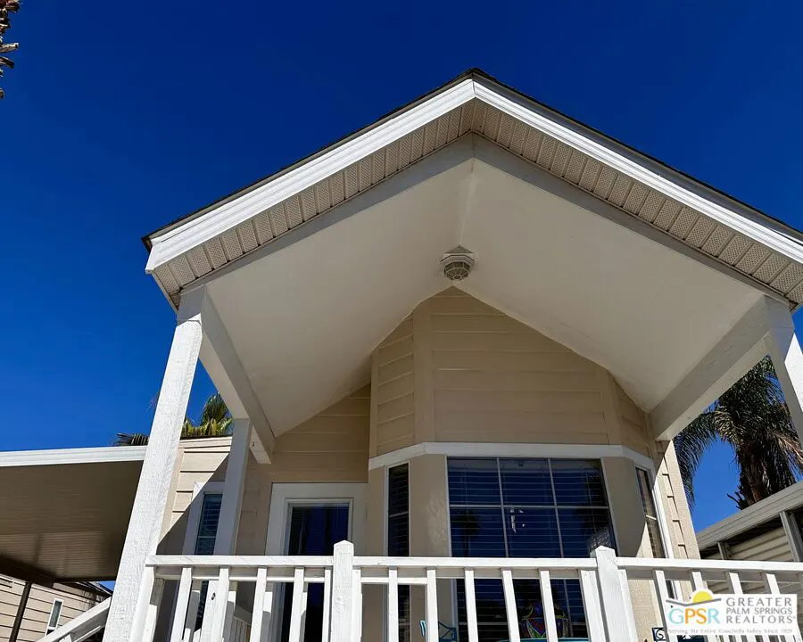 Exterior view of a beige house with white trim, a covered porch, and a white railing against a bright blue sky.