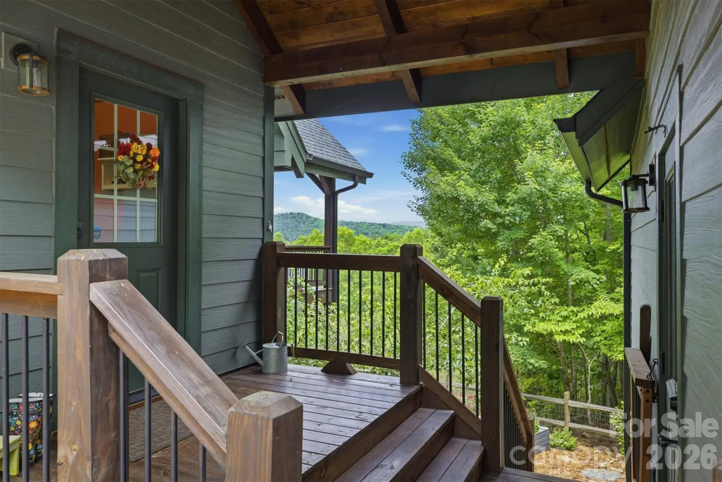 Exterior view of a gray house with a wooden porch, railing, and steps. A door with a wreath is visible, with a view of trees and mountains in the background.