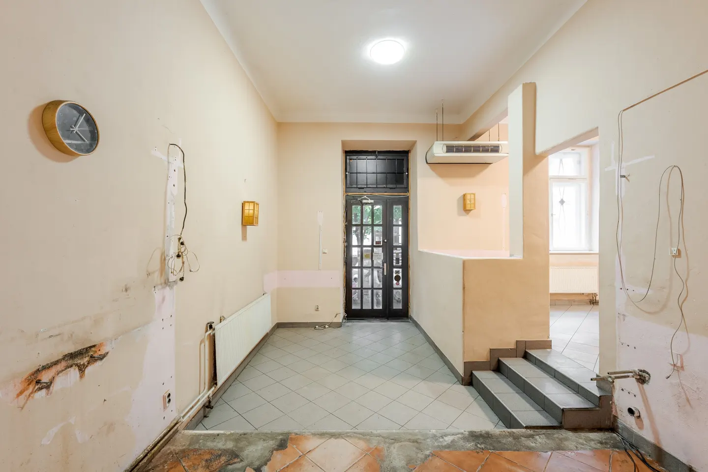 Interior view of a room under renovation with exposed wiring, a clock, and a black door leading outside. The floor is partially tiled.