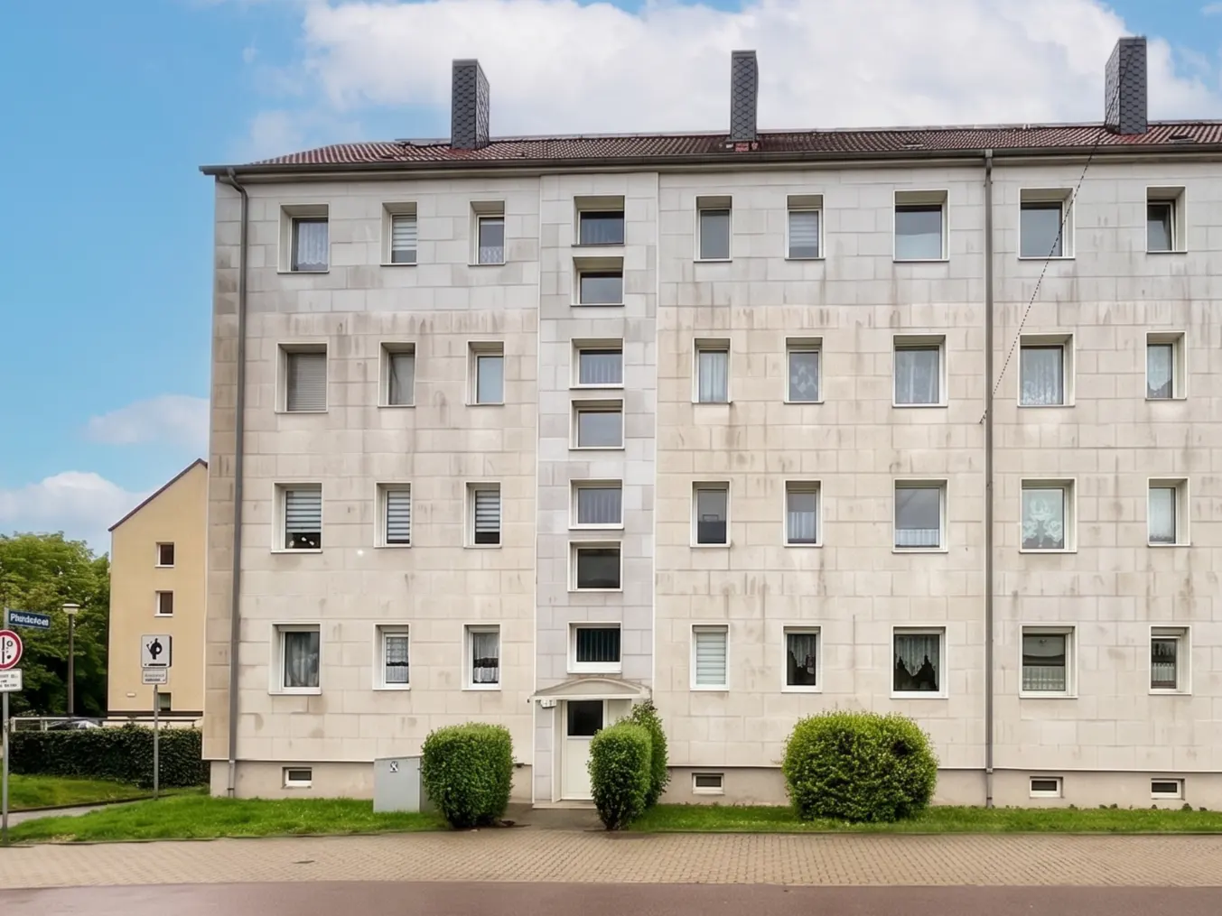 Exterior view of a five-story apartment building with a light gray facade and many windows. Shrubs line the base of the building.