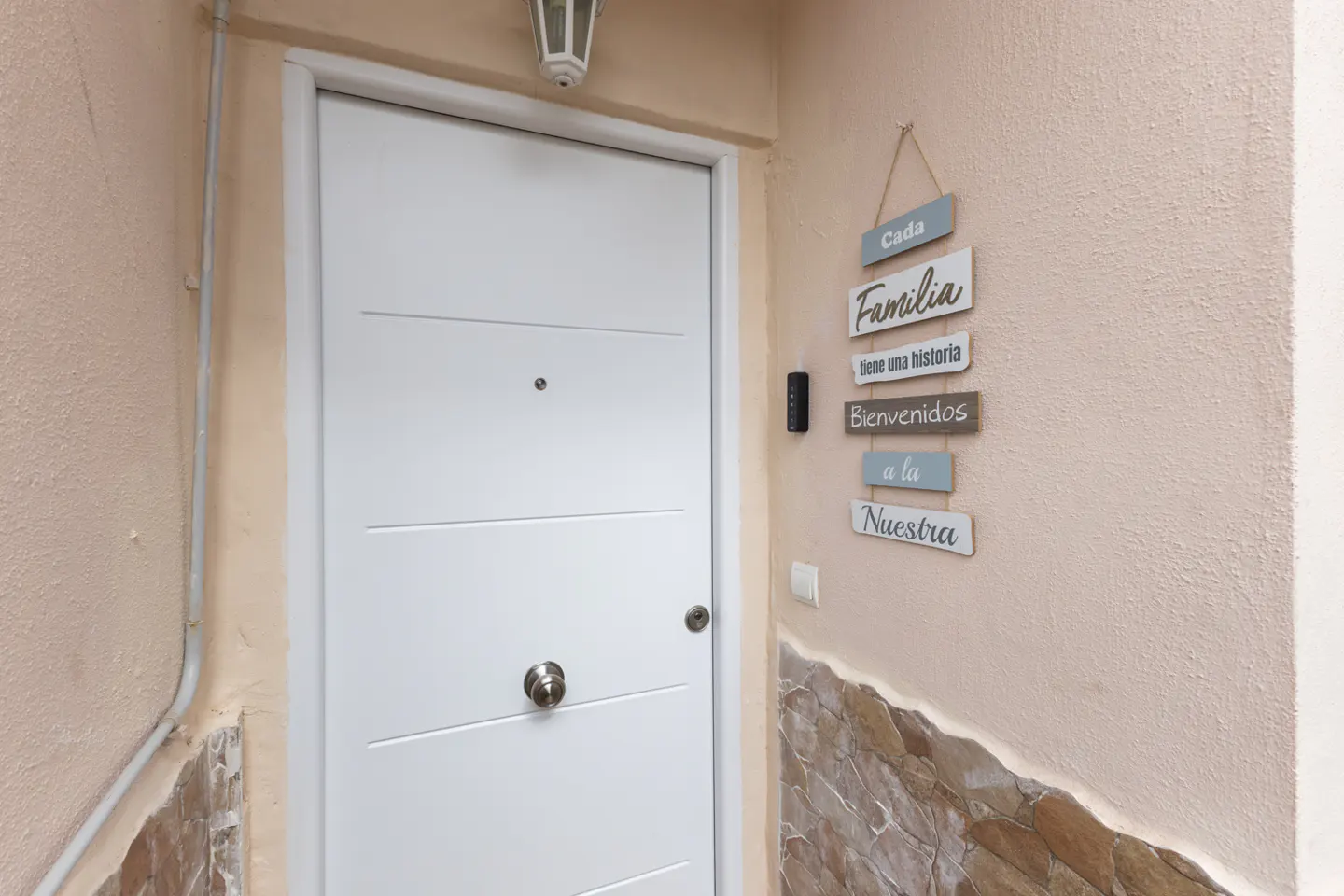 A white front door with a peephole and a silver knob, next to a sign that says "Cada Familia tiene una historia Bienvenidos a la Nuestra".