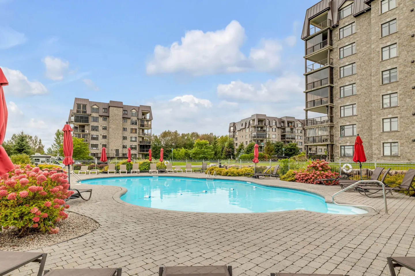 Outdoor pool area with lounge chairs, red umbrellas, and flowers. Stone buildings in the background under a blue sky with clouds.