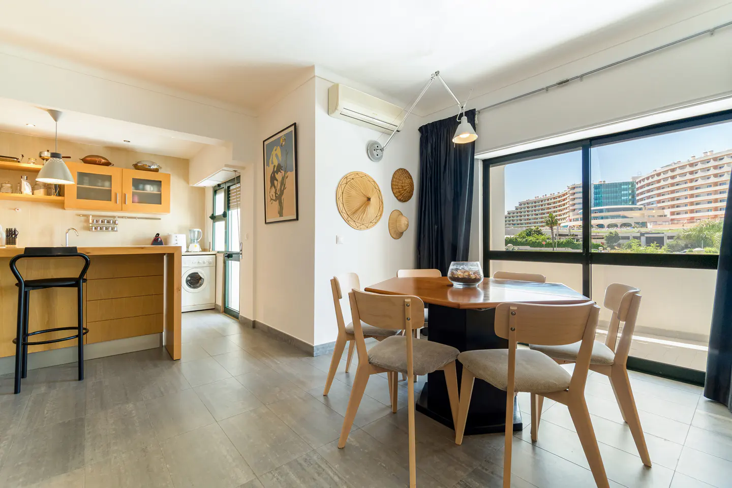 Dining area with a wooden table, four chairs, and wall-mounted straw hats, next to a kitchen with yellow cabinets.