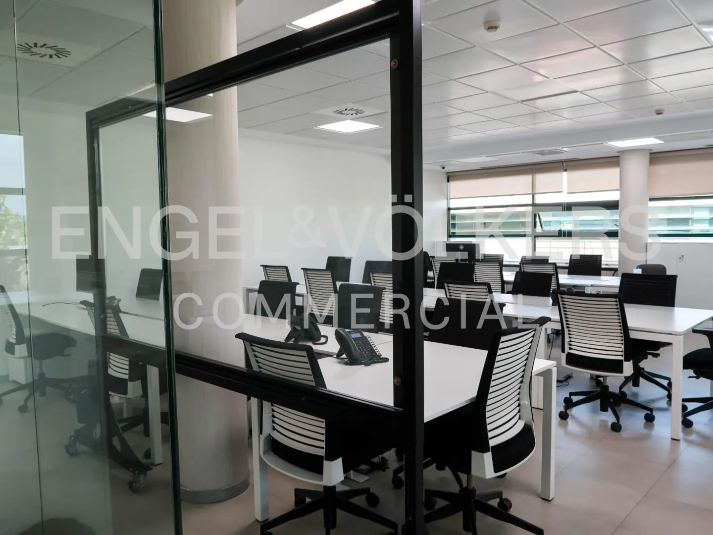 Office space with white desks and black chairs, seen through a glass wall with "Engel & Volkers Commercial" logo.