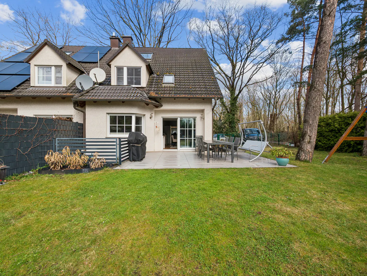 Backyard view of a two-story house with a patio, table, chairs, swing, grill, and green lawn. Solar panels on roof.