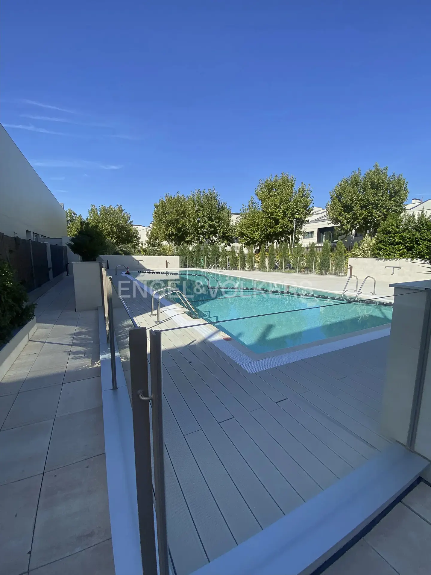 Outdoor pool with light blue water, surrounded by a gray deck and green trees under a clear blue sky. Engel & Volkers logo visible.