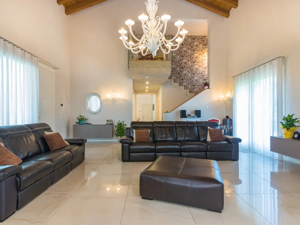 Bright living room with high ceilings, a white chandelier, and two black leather sofas. A staircase is visible in the background.