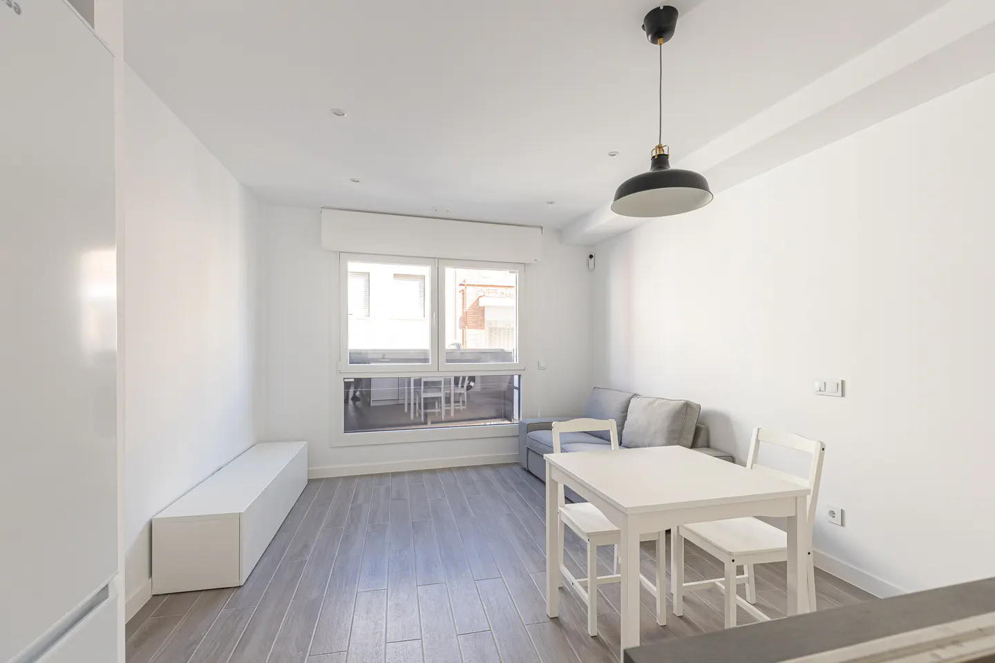 Minimalist living room with white walls, grey wood floors, a white dining set, grey sofa, and a black pendant light.