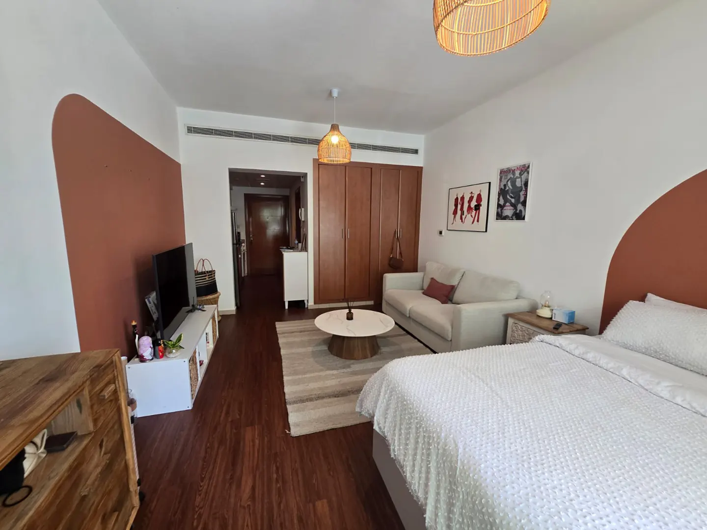 Bright studio apartment with white walls, brown accents, wood floors, and woven light fixtures. A white sofa and bed are visible.
