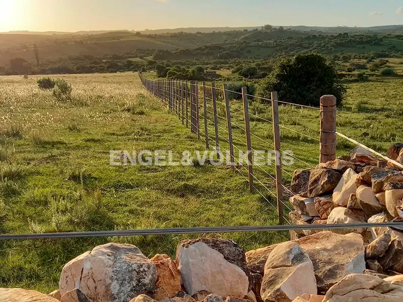 A wire fence stretches across a grassy field at sunset, with large rocks in the foreground and rolling hills in the distance.