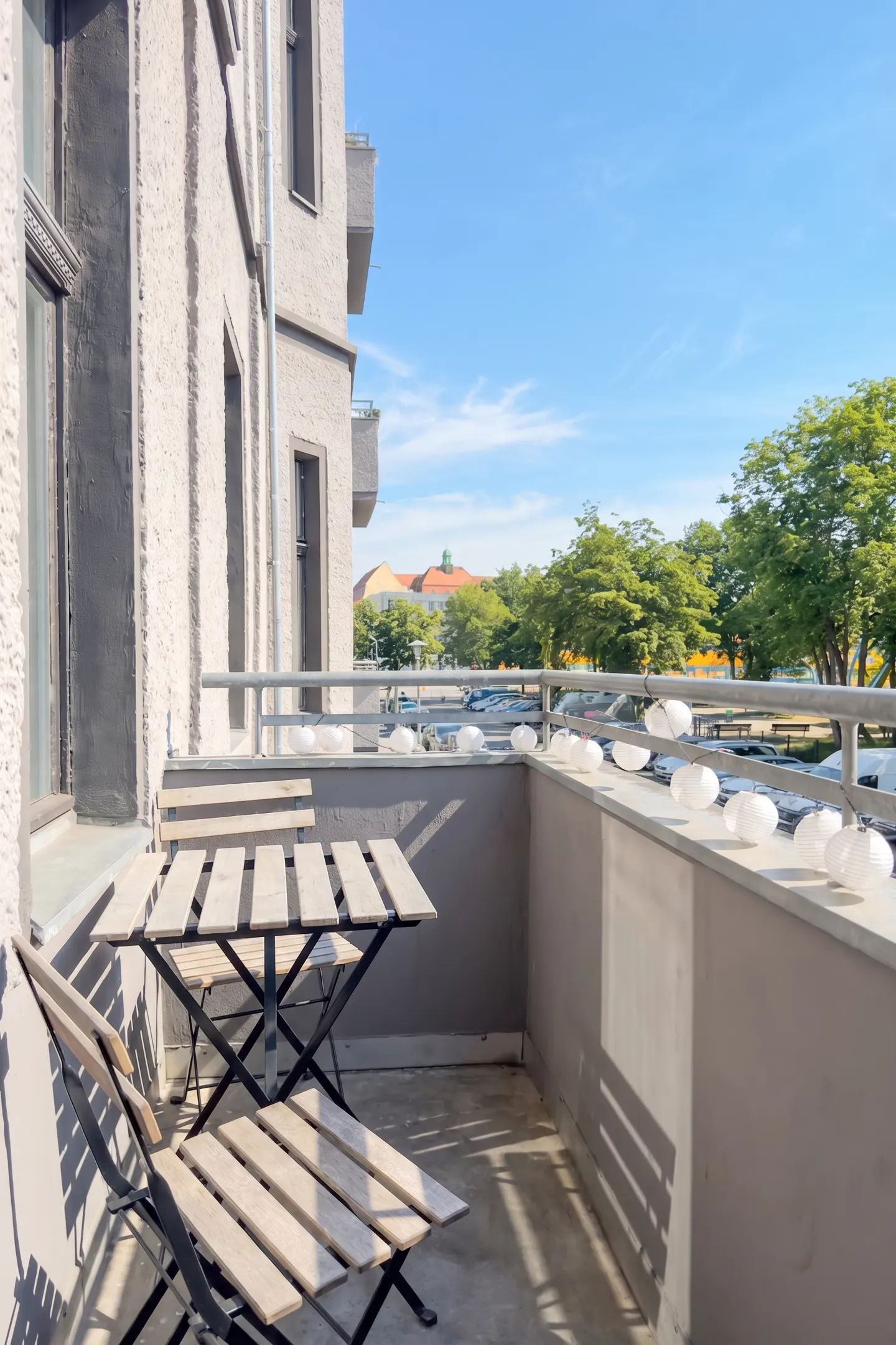 Balcony view with a wooden table and chairs, string lights, and a cityscape with trees under a blue sky.