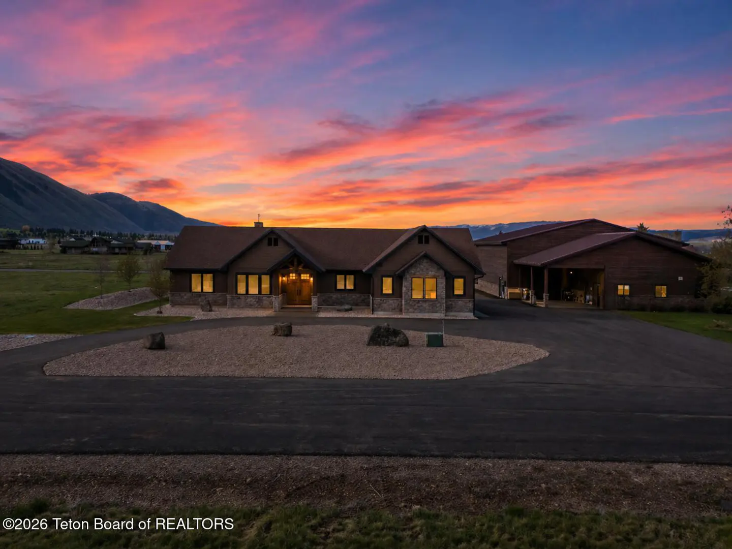 A brown house with lit windows sits on a gravel driveway under a colorful sunset sky.