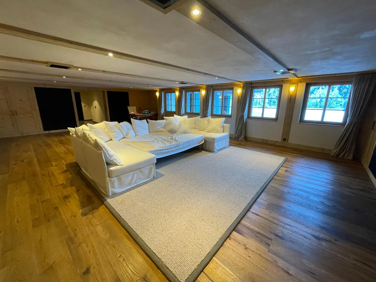 A spacious living room with a white sectional sofa, light carpet, and wood floors. Windows with curtains line the back wall.