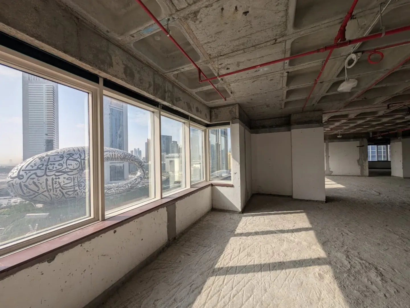 Unfinished office space with concrete floors and ceiling. Large windows offer a view of the Museum of the Future and city skyline.