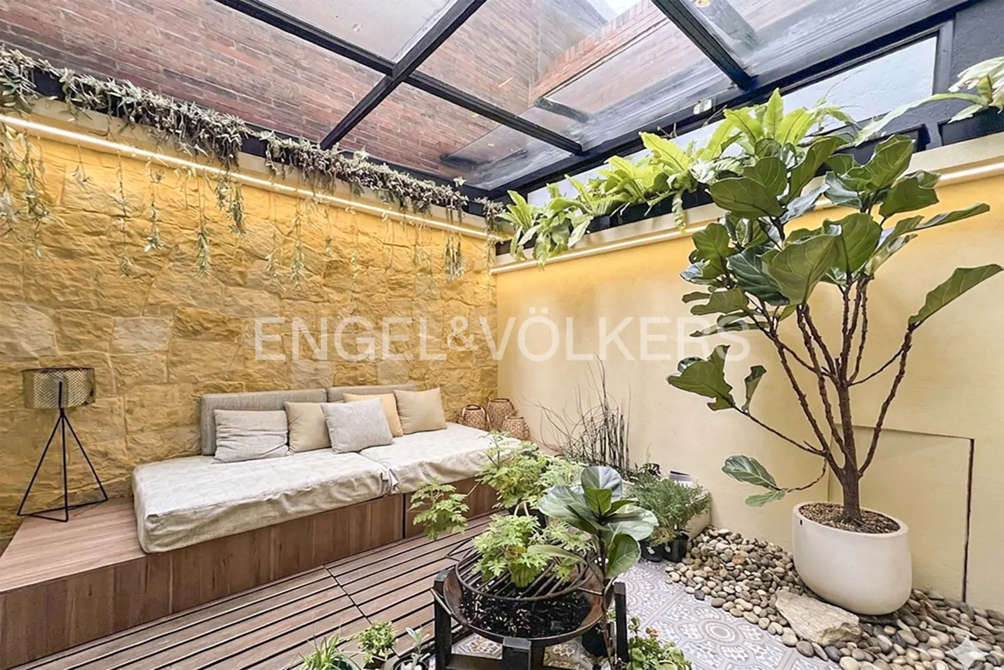 Indoor patio with stone wall, skylight, and wooden deck. A beige sofa with pillows sits against the wall. Plants and a small tree add greenery.