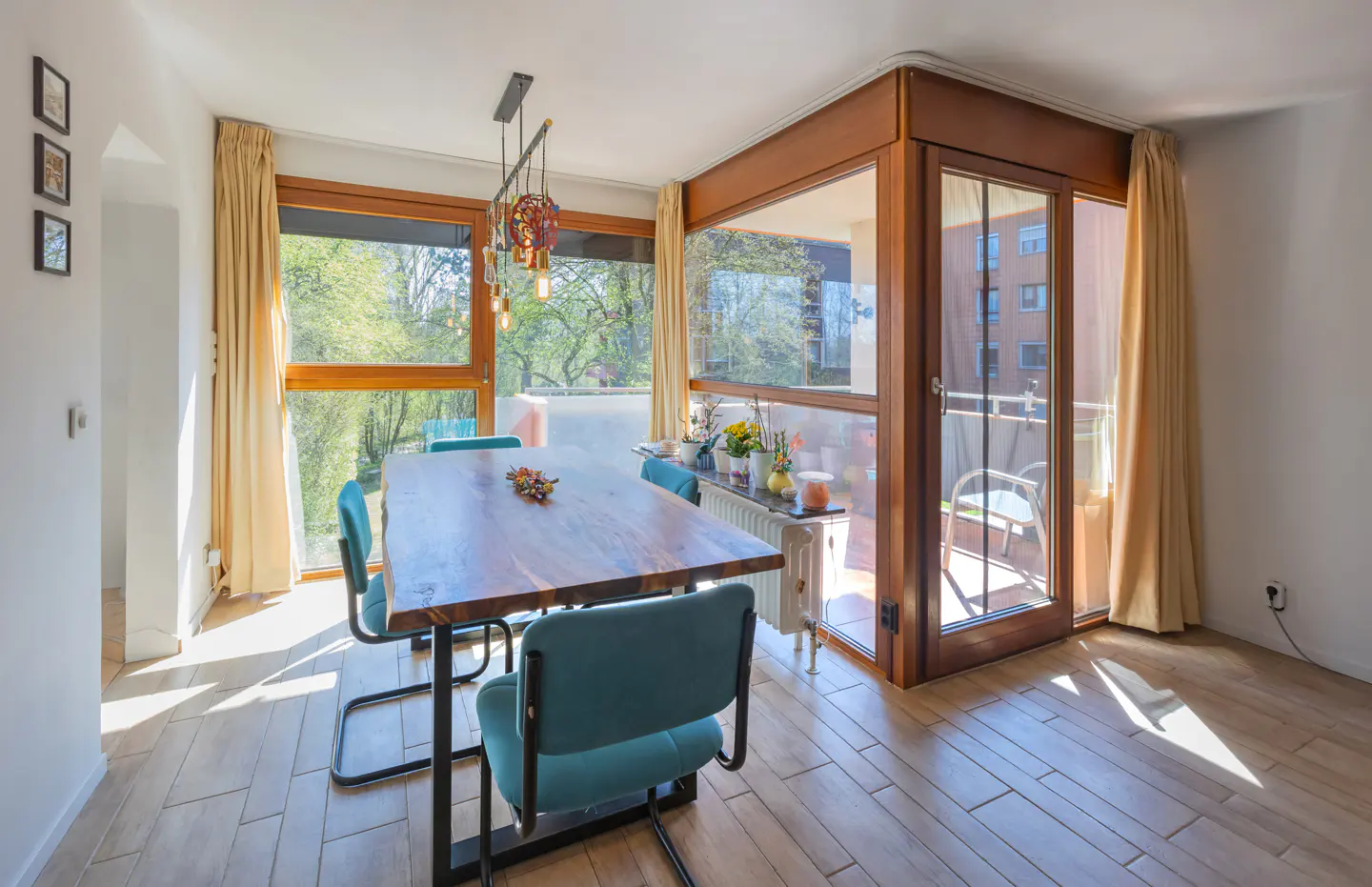 Dining room with a wood table, teal chairs, and a modern light fixture. Large windows overlook a green outdoor space and a balcony.