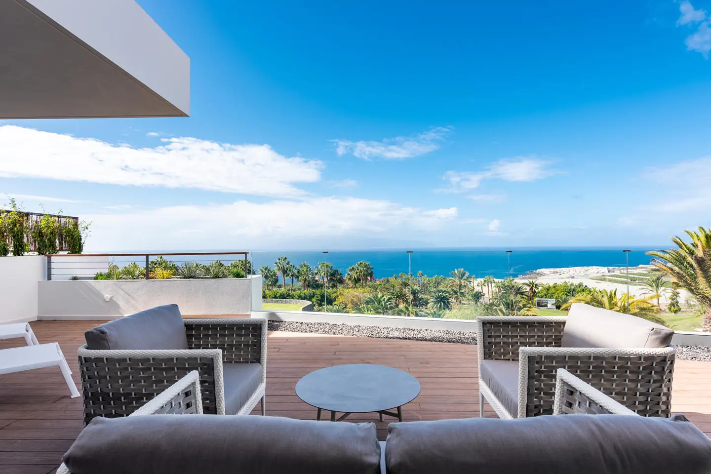 Outdoor patio with gray cushioned chairs and a round table overlooking the ocean and palm trees under a blue sky.