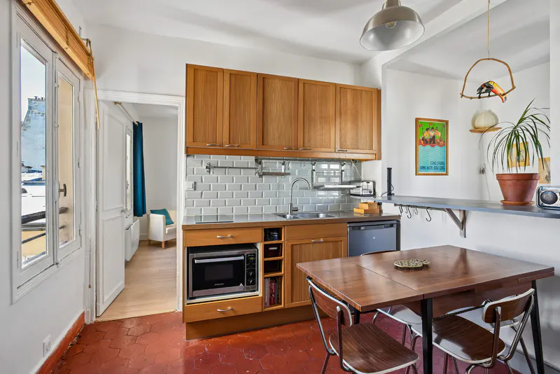A compact kitchen with wood cabinets, a gray tile backsplash, and a dining table with four chairs. A window and doorway lead to other rooms.