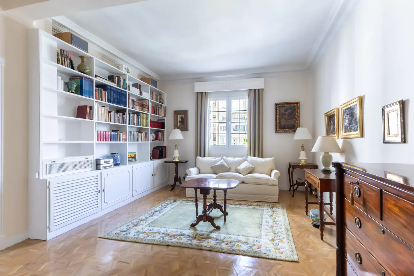 Bright living room with a white sofa, wooden floors, and a large white bookcase filled with books. A green rug sits under a dark wood coffee table.