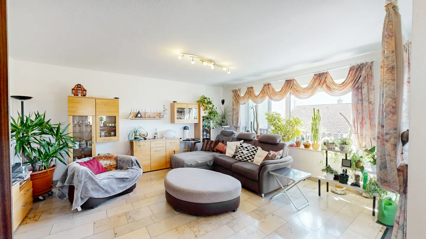 Bright living room with a brown sectional sofa, ottoman, and armchair. Large windows with floral curtains and many plants.