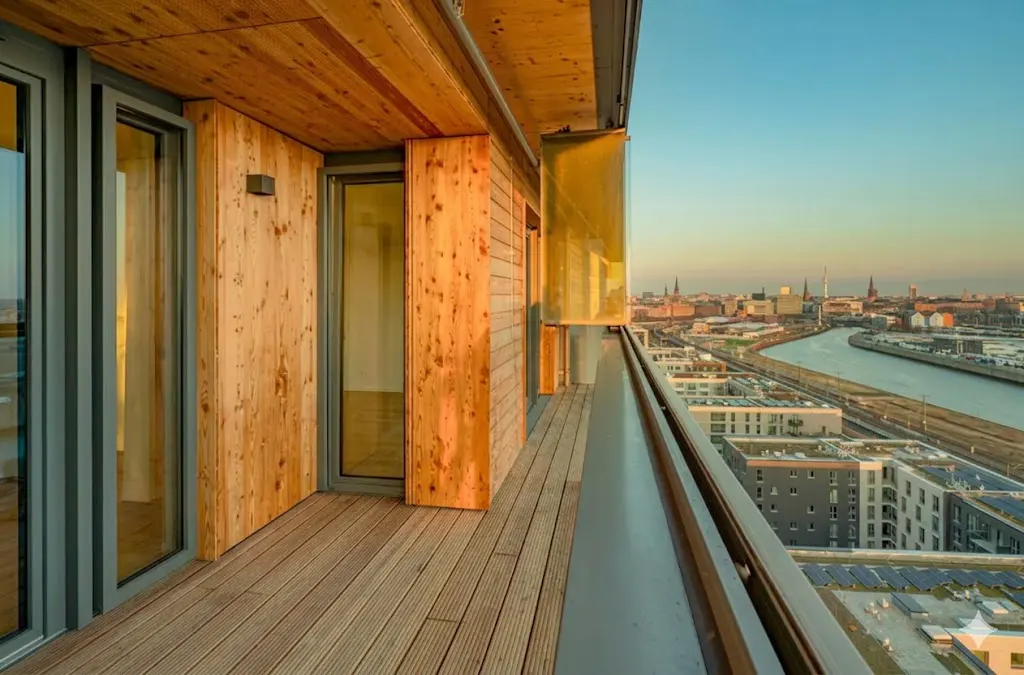Balcony view of a city skyline. Wood paneled walls and floor with glass doors. Metal railing overlooking buildings and a river.