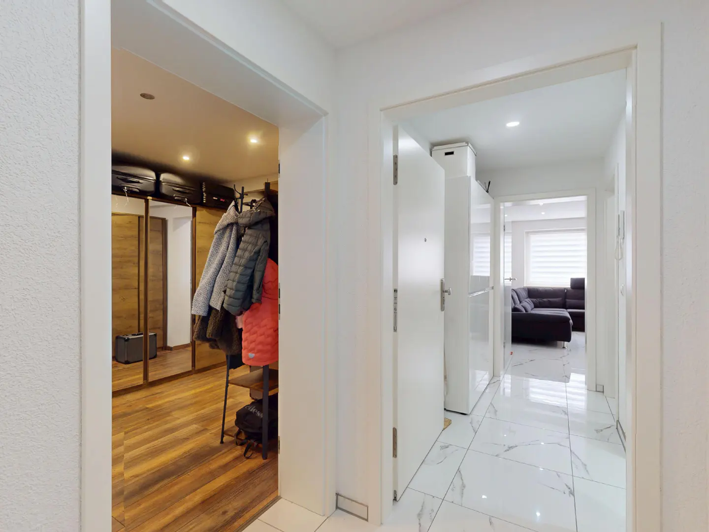 Hallway view with white walls, marble floors, and two open doorways. One leads to a closet with wooden floors and hanging coats, the other to a living room.